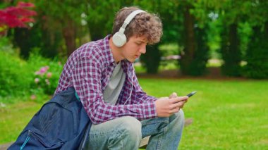 Kıvırcık çocuk kulaklıkla müzik dinliyor ve parkta otururken telefona bakıyor..