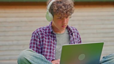 Kıvırcık çocuk kulaklıkla müzik dinliyor ve parkta otururken dizüstü bilgisayarla çalışıyor..
