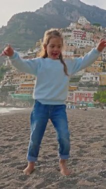  Positano sahilinde dans eden küçük tatlı kız gülüyor. Sahilden şehrin ana kısmına, dağdaki renkli evlere bakın. Positano, Amalfi Sahili, İtalya.