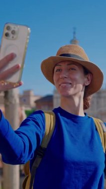  Şapkalı ve sırt çantalı mutlu bir turist kadın, Roma Forumu 'ndaki Antik Roma Sütunları' nın manzarasının arkaplanının önünde telefonuyla selfie çekiyor. İtalya.