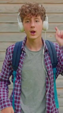 Dikey video. Genç, mutlu, kıvırcık kız kulaklıklarla dans ediyor, komik dans figürleriyle eğleniyor, sokaklarda özgürce yürüyor..