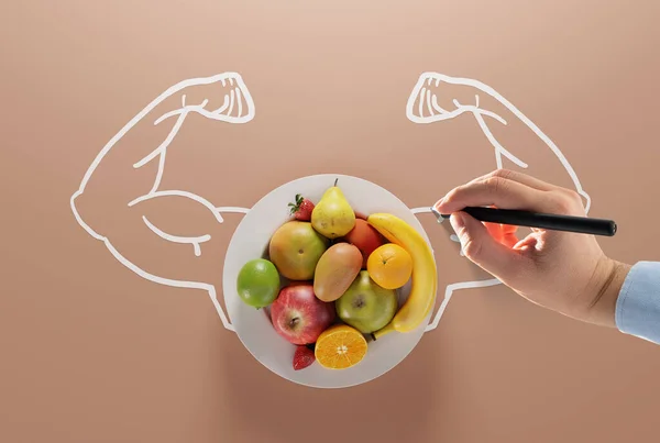 Hand paints muscles on the background of a plate with different fruits. The concept of healthy eating, fruits that give strength and essential food ingredients.
