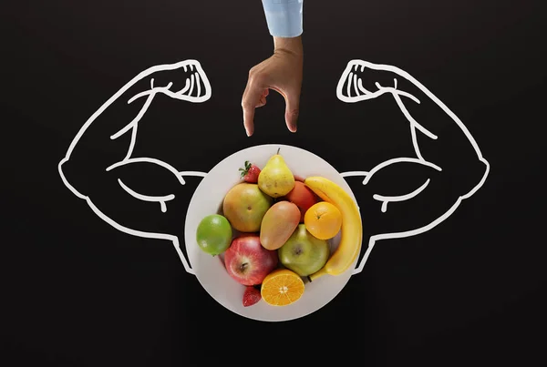 Muscles on the background of a plate and hand try to reach fruits. The concept of healthy eating, fruits that give strength and essential food ingredients.