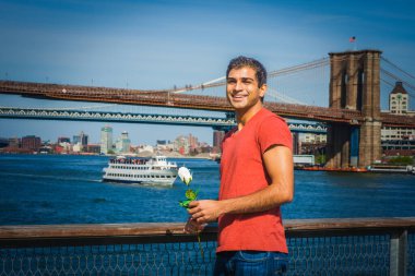 Doğu Hintli genç bir adam, kırmızı V yaka tişört giyiyor, kot pantolon, nehir kenarında duruyor, elinde beyaz bir gül, gülümsüyor ve seni bekliyor. Manhattan, Brooklyn köprüleri, arka planda tekne.