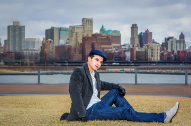 Şık giyinmiş bir adam çimenlerin üzerinde oturur, arka planda bulutlu bir gökyüzünün altında bir şehir silueti vardır. Koyu renk bir palto, kot pantolon ve düz bir şapka giyiyor. Sıradan ama modaya uygun bir hava yayıyor..