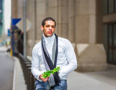 Eşarp ve ceket giyen genç bir adam elinde küçük bir buket çiçekle bir şehir caddesinde duruyor. Arkaplanda konuyu vurgulayan bulanık detaylarla şehir mimarisi yer alıyor.