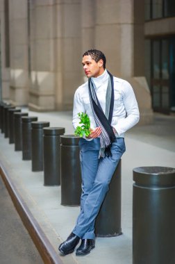 Bir insan, bir şehir caddesinde bir dizi silindirik direğe yaslanır ve elinde bir filiz yeşillik taşır. Beyaz gömlek, gri pantolon ve siyah eşarp giyince düşünceli görünüyorlar..