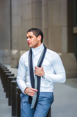 Modern bir binanın önünde ceketi ve atkısı olan bir adam duruyor. Rahatlamış ve kendinden emin görünüyor, bir eliyle atkıyı tutarken mesafeye bakıyor..