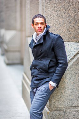 Şık giyimli bir adam şehir ortamında taş bir duvara yaslanır. Koyu renkli, çift göğüslü bir ceket ve açık gri bir pantolon giyiyor..