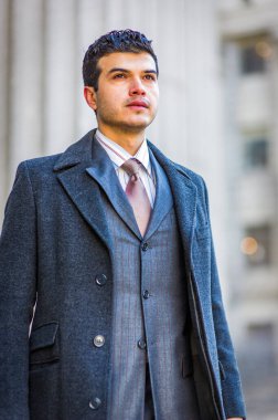 Şık gri paltolu ve takım elbiseli bir adam güvenli bir şekilde dışarıda dikilip mesafeye bakıyor. Arka planda bulanık mimari unsurlar şehir ortamını işaret ediyor