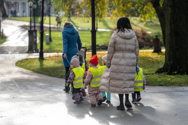Riga, Letonya - 4 Kasım 2022: Parkta yürüyüş için güvenlik yelekleri giymiş küçük çocuklu bir anaokulu öğretmeni