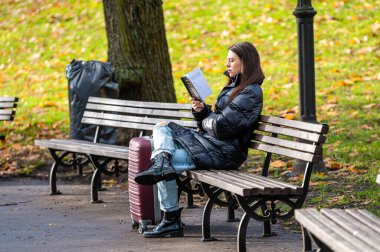 Riga, Letonya - 4 Kasım 2022: Genç bir kadın kitap okuyor ve sonbahar parkında bankta oturuyor. Sonbahar havası, boş zaman konsepti..