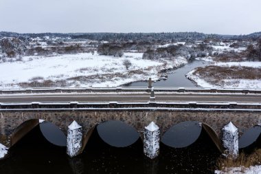 Karlı bir kış gününde Abava nehri üzerinde kemerleri olan eski bir taş köprü, Kandava, hava manzaralı.