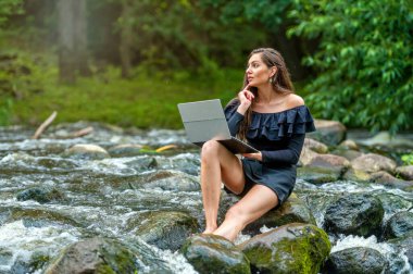 female freelancer sitting on a rock in the river and using a laptop, travel or vacation work concept