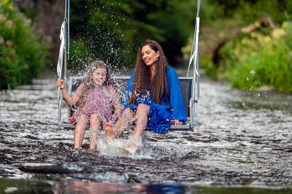 laughing woman and a young girl swing over a fast-flowing river, splashing the water with their feet