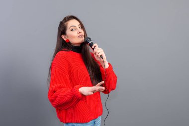 Genç, duygusal esmer, kırmızı kazak giymiş bir mikrofonla karaoke söylüyor ya da gri üzerine izole edilmiş bir konuşma yapıyor.