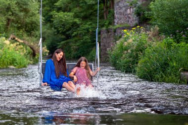 laughing woman and a young girl swing over a fast-flowing river, splashing the water with their feet