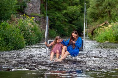 laughing woman and a young girl swing over a fast-flowing river, splashing the water with their feet