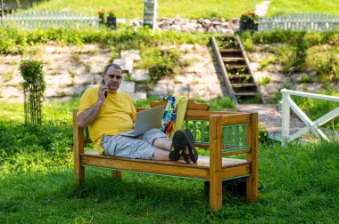 Orta yaşlı, dizüstü bilgisayarı ve akıllı telefonu olan bir adam bahçede çalışıyor, yeşil ofis kavramı.