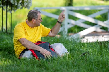 Sırt çantalı orta yaşlı bir adam güneşli bir günde çimlerin üzerinde oturur.
