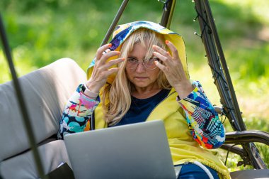 Bilgisayarlı ve belgeli olgun bir kadın bahçede sallanan koltukta çalışıyor, yeşil ofis konsepti.