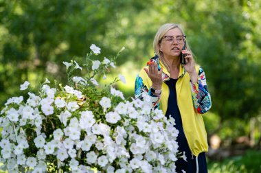 Parlak giyimli, sarışın, son sınıf öğrencisi, bahçede akıllı telefondan konuşan, online iletişim konsepti.