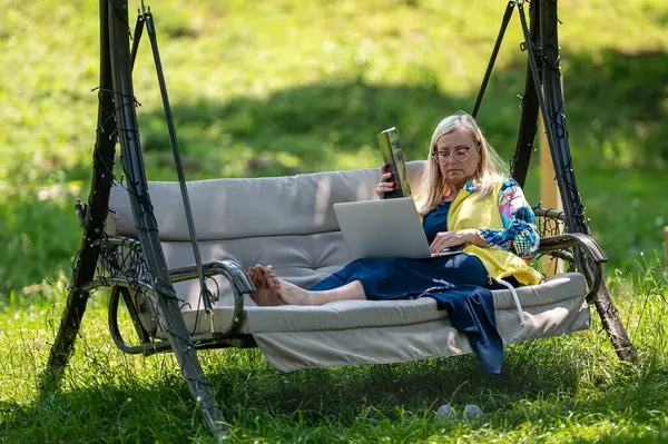 Bilgisayarlı ve belgeli olgun bir kadın bahçede sallanan koltukta çalışıyor, yeşil ofis konsepti.