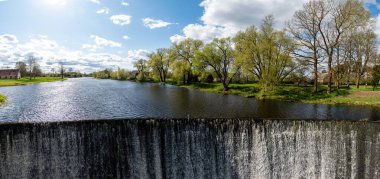 Nehir kenarındaki küçük bir köydeki yarı dairesel bir barajdan su akar. Bahar manzarası.