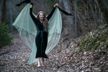 Bir kadın büyük kumaş kanatlarla ormanda yalınayak yürür. Suyun sığ derinliği