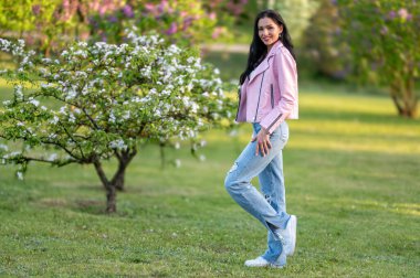 Pembe ceketli bir kadın, arka planda çiçek açan bir ağaçla parkta duruyor..