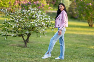 Pembe ceketli bir kadın parkta çiçek açan bir ağacın önünde poz veriyor. Suyun sığ derinliği