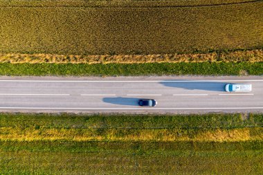 Arabalar yeşil tarlalar ve kahverengi tarım arazileriyle çevrili bir yol boyunca sürüyorlar..