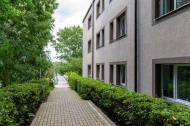 A peaceful walkway is bordered by lush greenery and modern buildings on a cloudy day. clipart