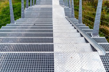 Güneşli bir günde yemyeşil bitki örtüsüyle çevrili çelik ızgaradan yapılmış yaya köprüsü basamakları..