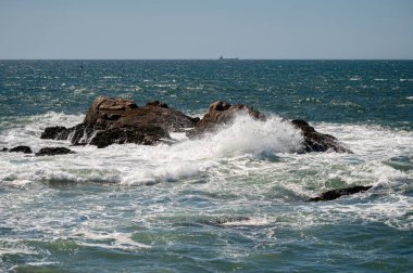 Güçlü okyanus dalgaları açık bir gökyüzünün altında sahil boyunca uzanan büyük kayalarla çarpışır..