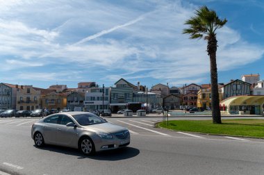 Costa Nova, Portekiz - 11 Eylül 2024: Parlak bir gökyüzü altında büyüleyici binaların ve palmiye ağaçlarının yanından geçen gümüş bir araç.