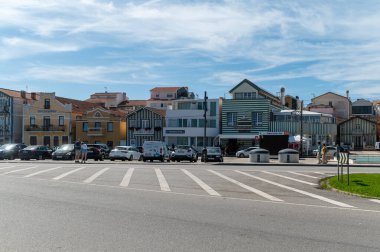 Costa Nova, Portekiz - 11 Eylül 2024: Ziyaretçiler mavi gökyüzünün altındaki büyüleyici bir sahil kasabasında park halindeki arabaların yanında geziniyorlar.