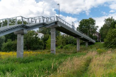 A modern pedestrian bridge spans lush greenery in an urban setting on a sunny day clipart