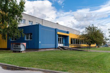 Talsi, Letonya - 1 Eylül 2024: Parlak renkli, yeşil çimenli, bulutların çerçevelediği endüstriyel bir binanın dışı.