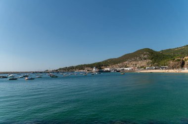 Sesimbra, Portekiz - 10 Eylül 2024: Sakin sularda dinlenen teknelerin sakin manzarası kumlu bir sahil ve çevreleyen tepeler.