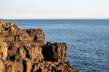Engebeli kayalar gün batımında berrak bir gökyüzünün altında sakin okyanusu özetliyor..