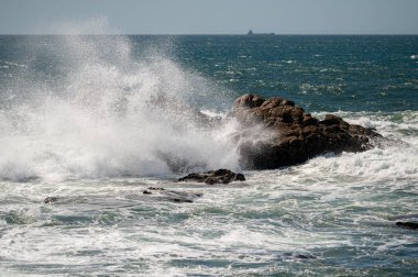 Dalgalar açık denizde parlak güneş ışığı altında kayalık kıyılara çarpıyor.