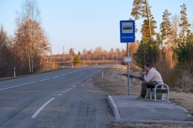 Bir adam otobüs durağında tek başına oturuyor, etrafı ağaçlarla çevrili, şafak vakti ulaşımı bekliyor..