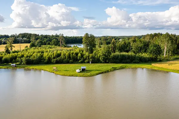 Ağaçlarla çevrili sakin bir göl manzarası ve mavi gökyüzünün altındaki açık alanlar..