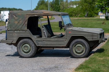 Askeri tarzda bir cip, yeşilliklerle çevrili çakıllı bir yüzeyde güneşin altında öylece durur..