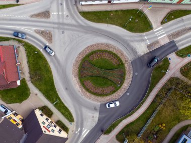 Çevresindeki yeşil peyzaj ve binalarla çevrili bir kavşakta araçlar geziniyor..