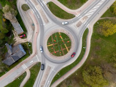 Arabalar, güneşli bir günde yeşil alanlar ve patikalarla çevrili bir kavşakta yol alırlar..