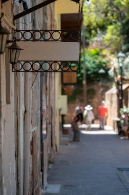 Dar bir caddenin yanındaki duvarda asılı boş tabela ve yakınlarda keşifler yapan yayalar var..