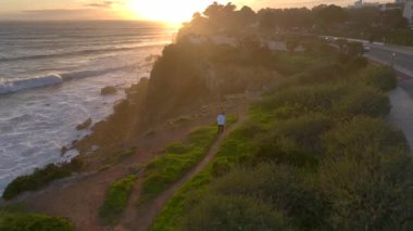 Okyanusun kıyısındaki uçurumun kenarında koşan bir adam var. Genel bakış açısı, Cascais, Portekiz 'de gün batımı zamanı. Hava Aracı Görünümü. Yüksek kalite 4k görüntü