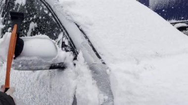 Cleaning car windshield and rearview mirror from snow with brush and scraper tool during heavy snowfall in Winter. Car covered in snow.
